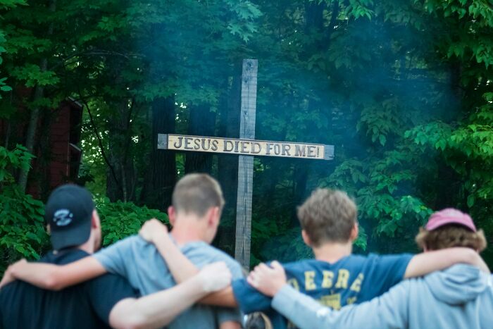 Four young men with arms around each other facing a wooden cross in a wooded area, relating to former cult members.