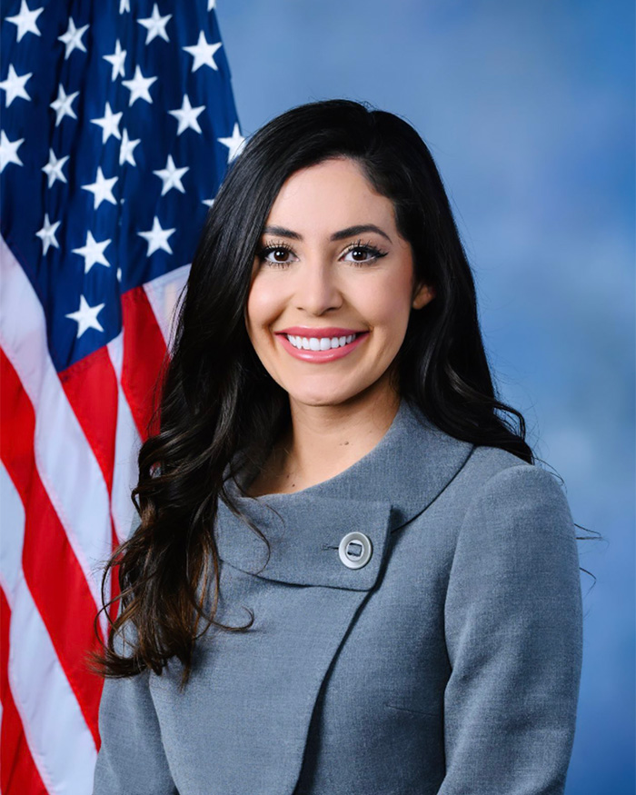 Erika Kirk smiling in a gray coat with the American flag in the background amid custody speculation news.
