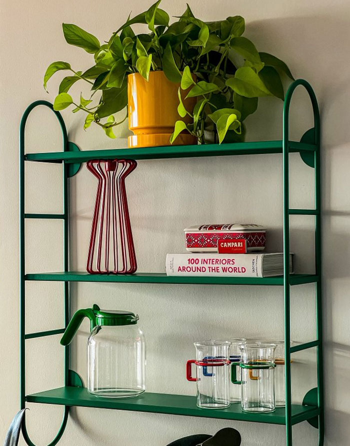Green metal wall shelf with home decor items including a plant, books, and glassware for cool interior design ideas.