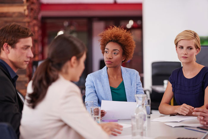 A young woman shares her cancer surgery story during a serious discussion with coworkers at a meeting table.