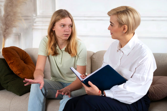 Two women having a serious conversation on a couch, one sharing her cancer surgery story with the other listening intently.