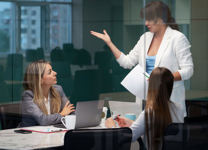 Three professional women in a meeting room discussing, highlighting a 28-year-old sharing cancer surgery story.