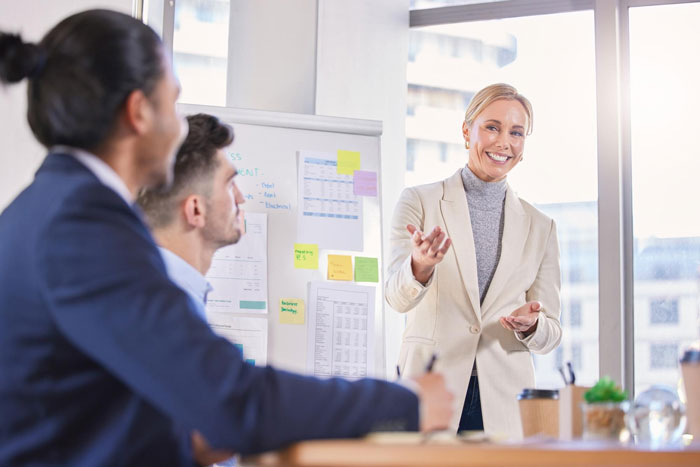 Office Karen belittles coworker during meeting while others listen in a modern bright office with charts on a whiteboard.