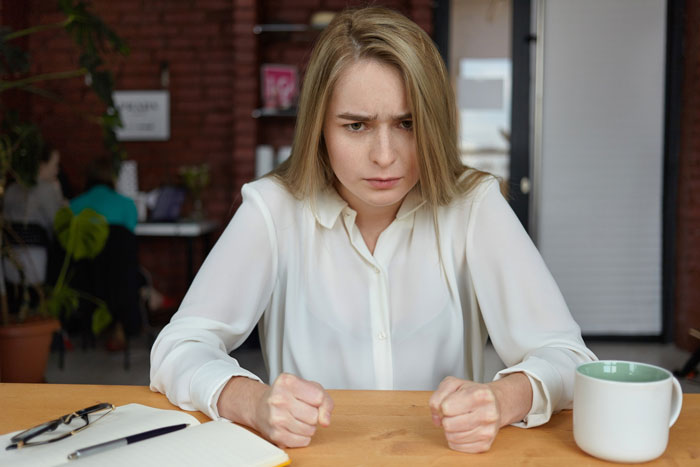 Frustrated office woman sitting at desk with clenched fists, experiencing tension in a workplace Karen conflict.