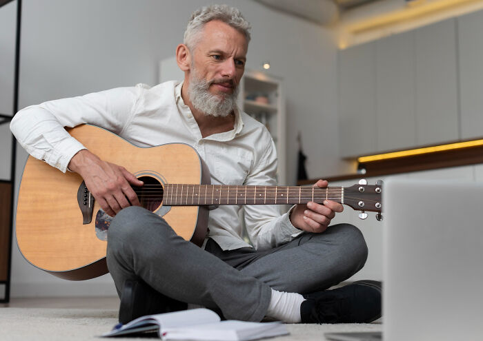 Middle-aged man playing acoustic guitar on floor near laptop, notebook open, pondering high school secrets.