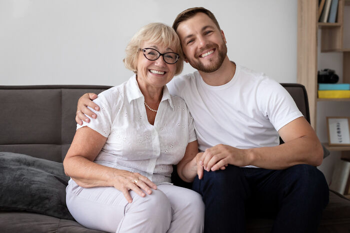 Young man and elderly woman smiling on couch, holding hands, hinting at shared high school secrets discovered later