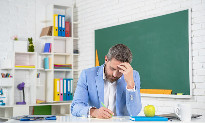 High school secrets: stressed male teacher in classroom writing at desk with chalkboard, apple, books, and mug