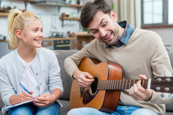 Man playing acoustic guitar for smiling teen taking notes, suggesting high school secrets and late discoveries