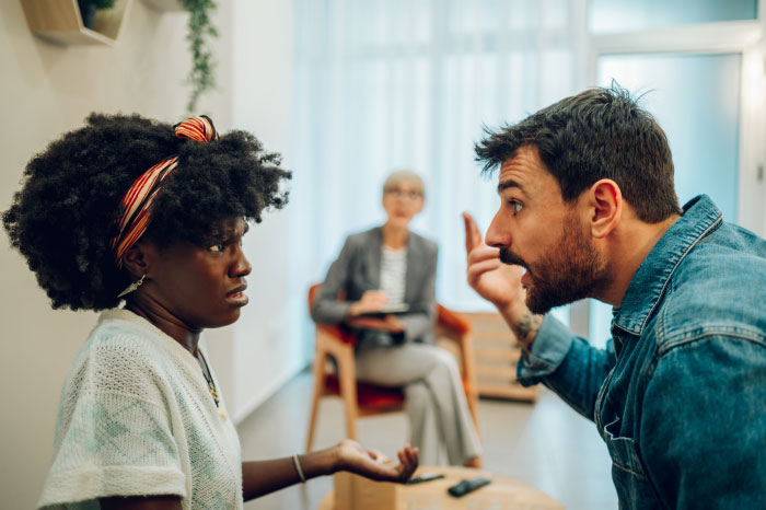 A tense family argument as a man verbally attacks a woman while an older woman observes, highlighting family conflict and poisoning issues.