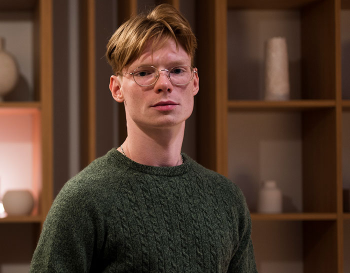 Young man with glasses wearing green sweater, standing indoors with calm expression, showcasing sharp intelligence vibe.