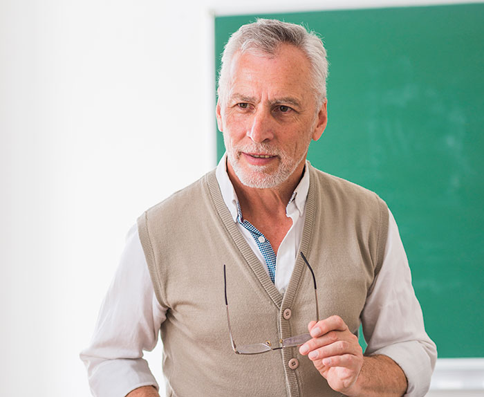 Older male teacher holding glasses by a chalkboard, demonstrating intelligence so sharp it almost feels like brain spaghetti.