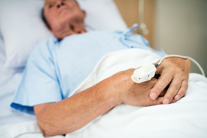 Hospital workers view elderly patient lying in bed with pulse oximeter on finger, hands clasped under blanket