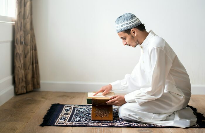 Young man in traditional clothing reading a book on a prayer rug, illustrating cultural norms and differences worldwide.