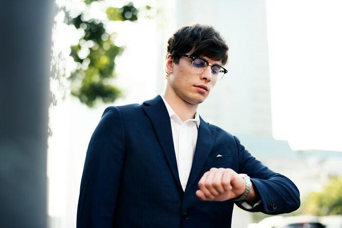 Young man in a blue suit and glasses checking his watch outdoors, illustrating cultural differences in behavior worldwide.
