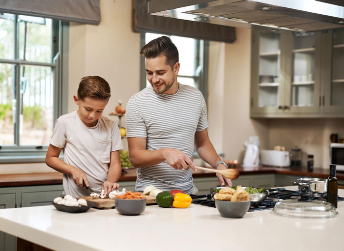 Dad and brother cooking food together in a bright kitchen with fresh vegetables and ingredients on the counter.