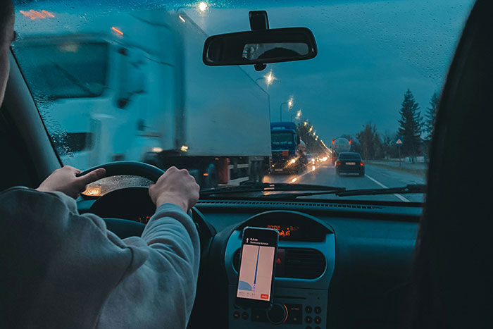Driver holding steering wheel on rainy road at dusk with large trucks passing by, showing dangerous driving conditions.