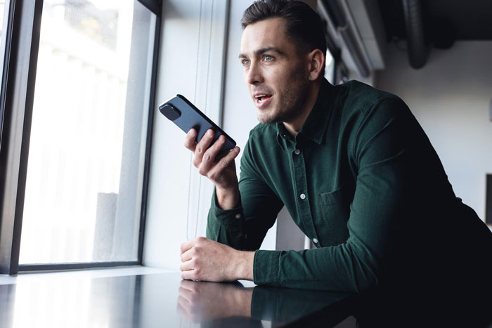 Man holding smartphone near window, appearing surprised while talking inside a modern cafe or bar setting.
