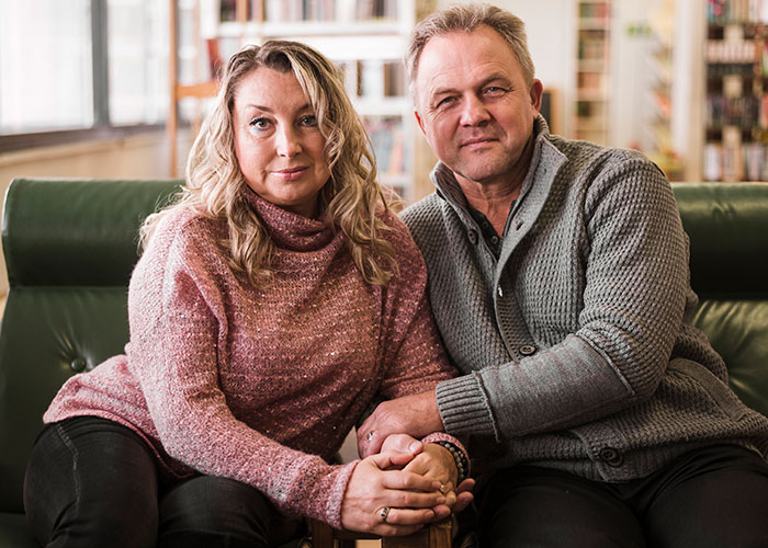 Middle-aged woman and man sitting closely on a couch, holding hands, considering a DNA test with mixed emotions. Middle-aged woman and man sitting closely on a couch, holding hands, considering a DNA test with mixed emotions.