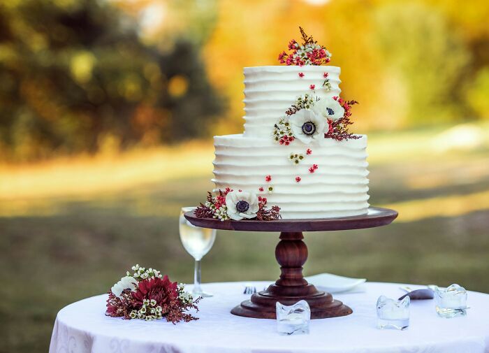 Two-tier white wedding cake with floral decorations on wooden stand at outdoor wedding moments.