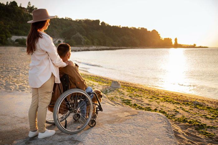 Woman standing behind disabled son in wheelchair at beach during sunset reflecting on their challenging family life.