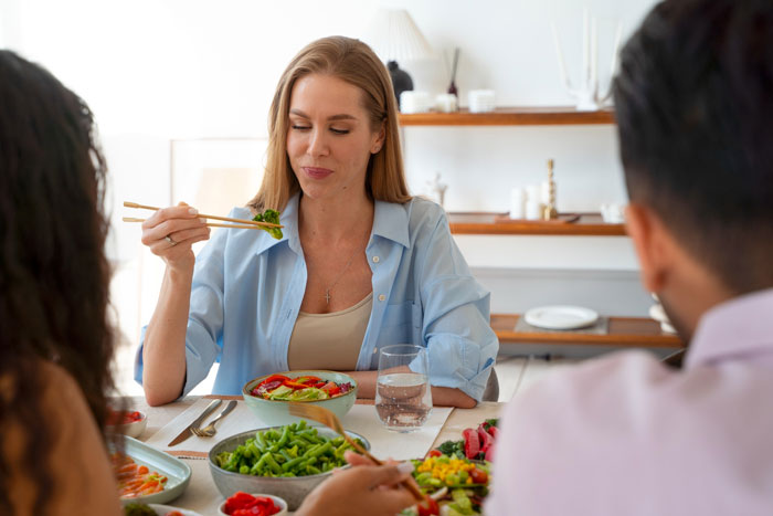 Woman eating at a family dinner while others, including a man, sit around the table in a bright room.
