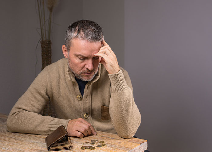 Man in a beige sweater looking worried about finances, counting coins on a wooden table, representing financial problems.