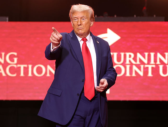 Former President Trump in a navy suit and red tie pointing, highlighting changes in Trump&rsquo;s hair color by a professional stylist.