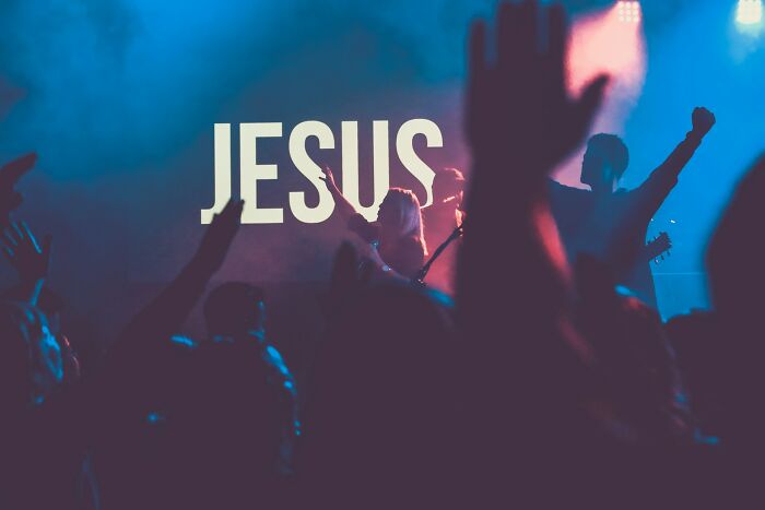 Crowd at a concert with hands raised, illuminated by blue and red lights, highlighting the word Jesus on screen related to former cult members.