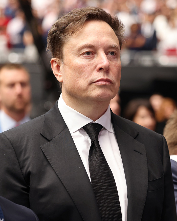 Elon Musk in a black suit and tie at a formal event, making a dramatic announcement about his 13th child.