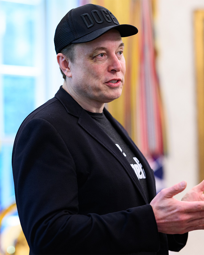 Elon Musk wearing a black hat and blazer speaking indoors, related to announcement about 13th child with influencer.