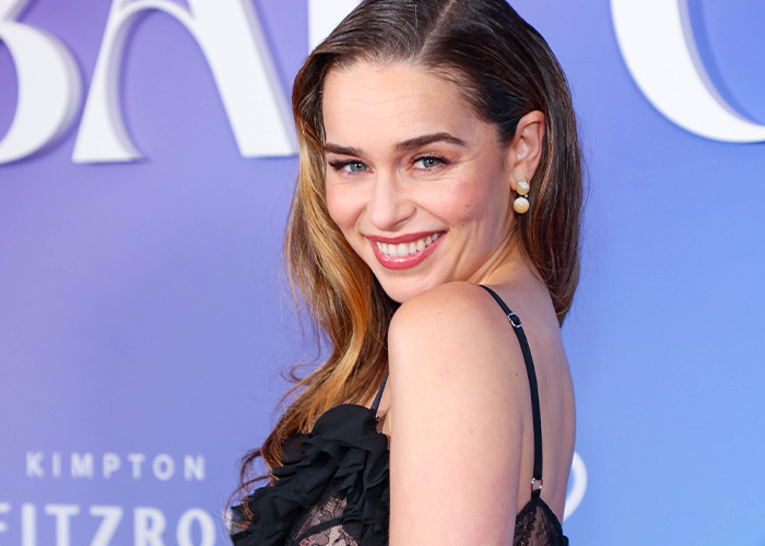 Emilia Clarke smiling at a formal event, wearing black lace dress and pearl earrings, with styled hair and natural makeup