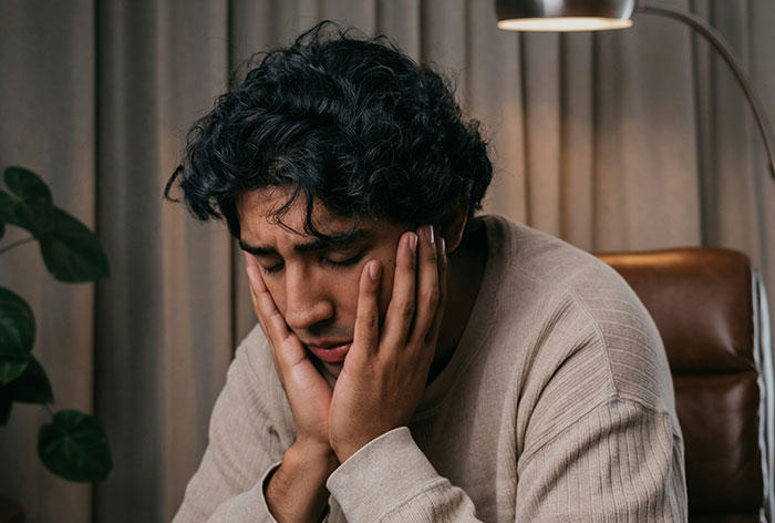 Young man with curly hair looking distressed, sitting indoors with hands on his face, reflecting on an unexpected family discovery.