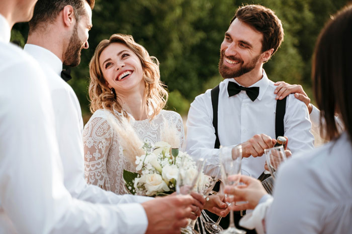 Bride and groom celebrating engagement hold with friends outdoors, smiling and raising glasses in a joyful moment.