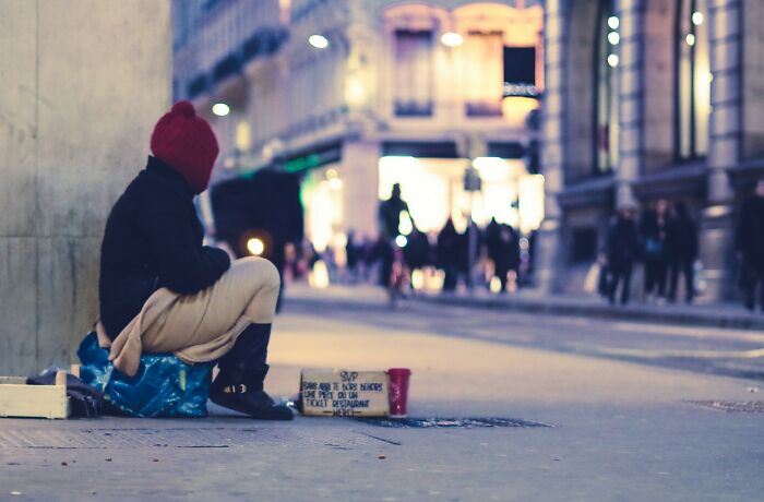 Person sitting on sidewalk near wall with a sign and cup, urban street scene in the evening with blurred pedestrians.