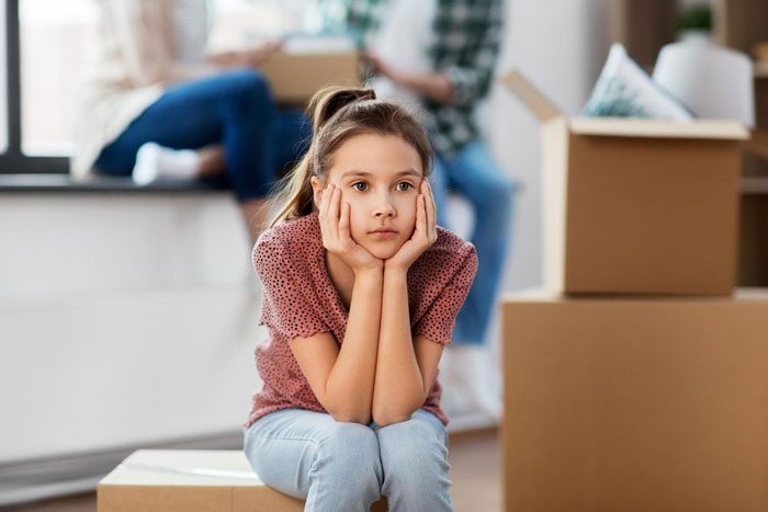 Bratty teen girl sitting on a box looking frustrated with moving boxes and adults in the background.