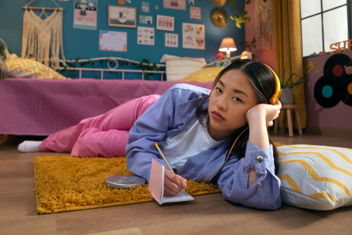 Teen girl lying on bedroom floor with headphones, writing in notebook surrounded by colorful decorations and a messy room.