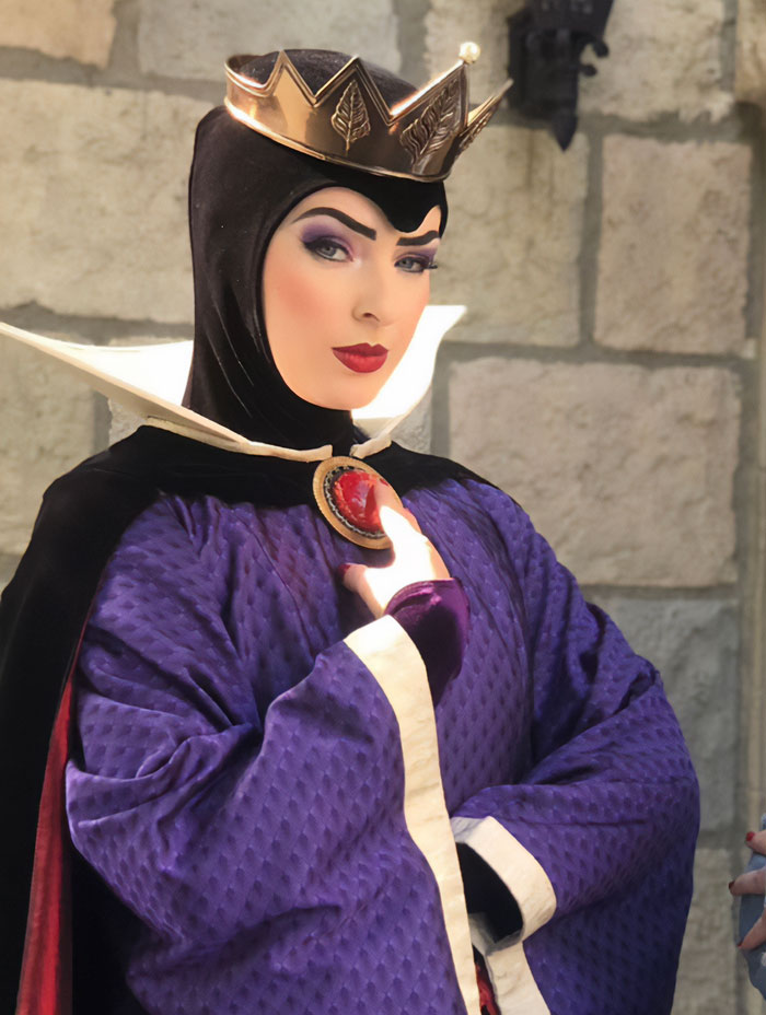 Evil Queen in detailed costume with crown and makeup, posing in front of a stone wall at Disneyland.