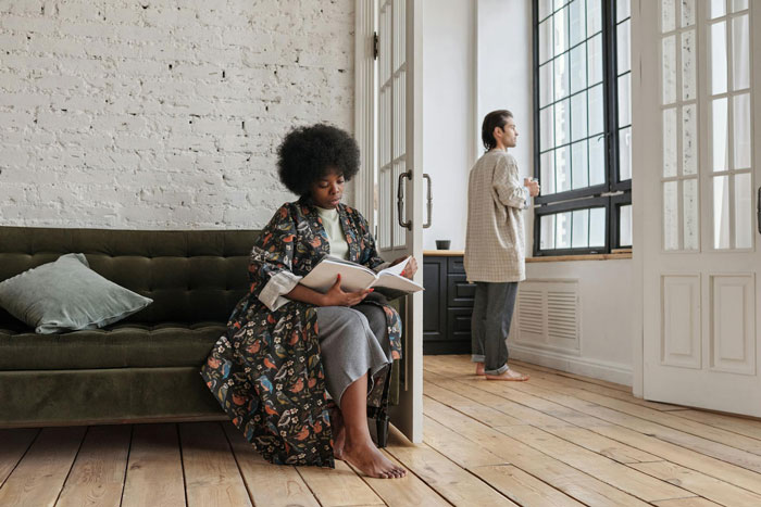 A woman reading on a sofa while her ex stands by the window in a tense ex house drama relationship scene.