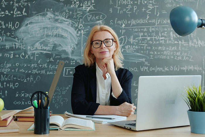 Woman expert with glasses sitting by laptop in front of a chalkboard covered with complex mathematical formulas.