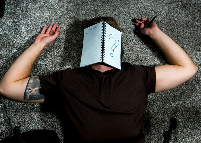 Person lying on carpet with notebook covering face showing a large question mark, representing overused slang words to banish. Person lying on carpet with notebook covering face showing a large question mark, representing overused slang words to banish.