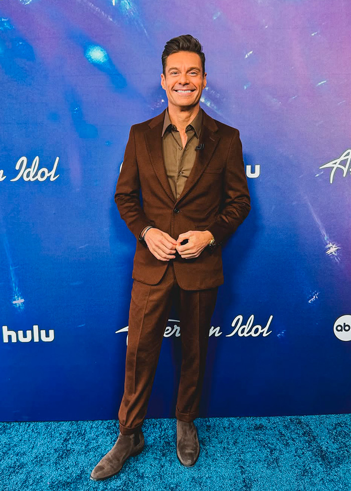 Ryan Seacrest standing on a blue carpet wearing a brown suit, showing an extremely skinny look of his face.