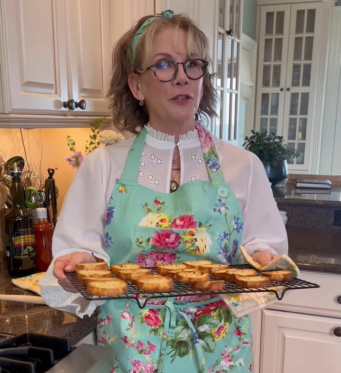 Melissa Gilbert in a kitchen wearing a floral apron holding a tray of toasted bread during a fridge tour video.