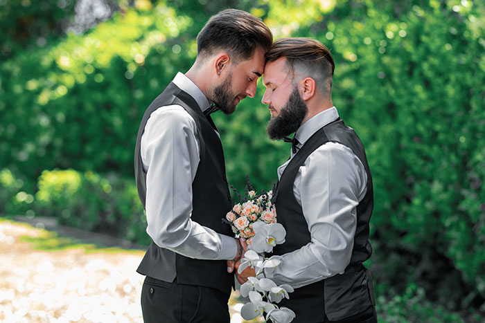 Two men in matching vests share a tender moment outdoors, representing a gay son's extravagant wedding celebration.