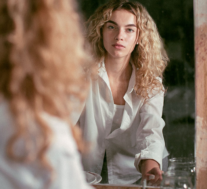 Young woman with curly hair looking thoughtfully at her reflection, highlighting women’s bodies and self-awareness.