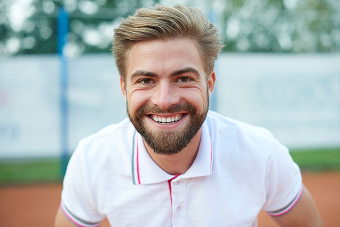 Smiling young man with beard wearing a white polo shirt, outdoors on a sports court, representing women’s body awareness.
