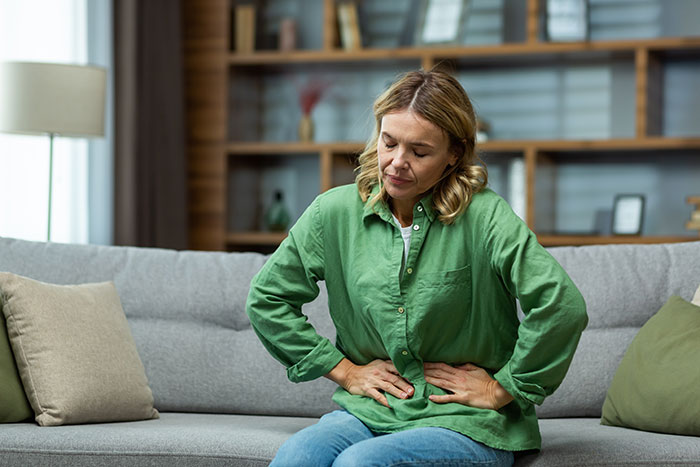 Middle-aged woman sitting on a couch holding her abdomen, reflecting concerns about women's bodies and health issues.