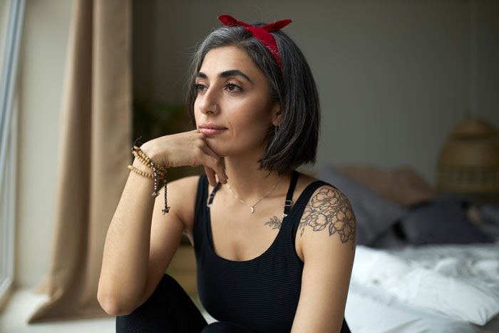 Woman with floral tattoo on shoulder wearing a red headband, looking thoughtful indoors by a window in casual wear. Woman with floral tattoo on shoulder wearing a red headband, looking thoughtful indoors by a window in casual wear.