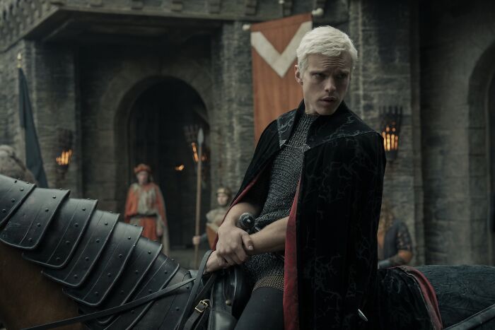 Actor portraying a Targaryen character in A Knight Of The Seven Kingdoms wearing a black cloak and holding a sword on horseback.