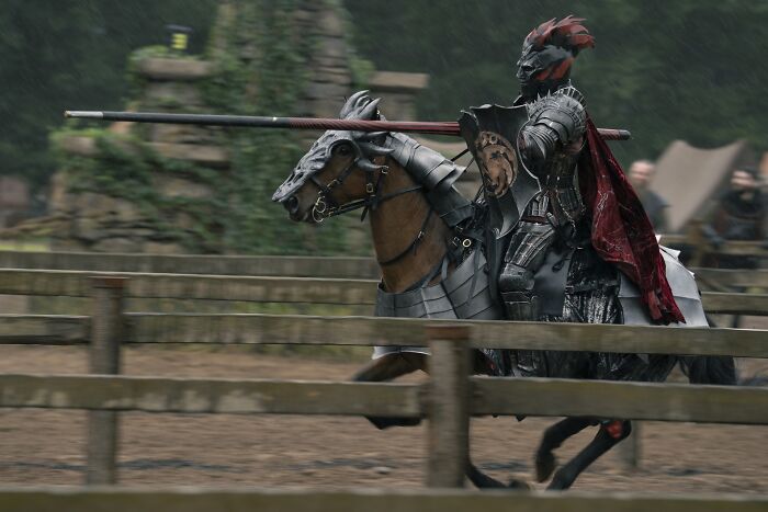 A knight in black armor with a red cape jousting on a horse during a medieval tournament scene. A knight in black armor with a red cape jousting on a horse during a medieval tournament scene.