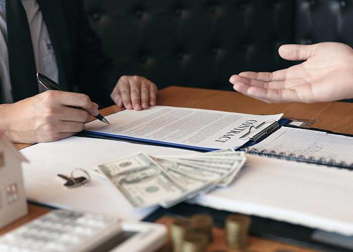 Person signing a real estate contract at a desk with money, keys, and a small house model, illustrating buying a house.